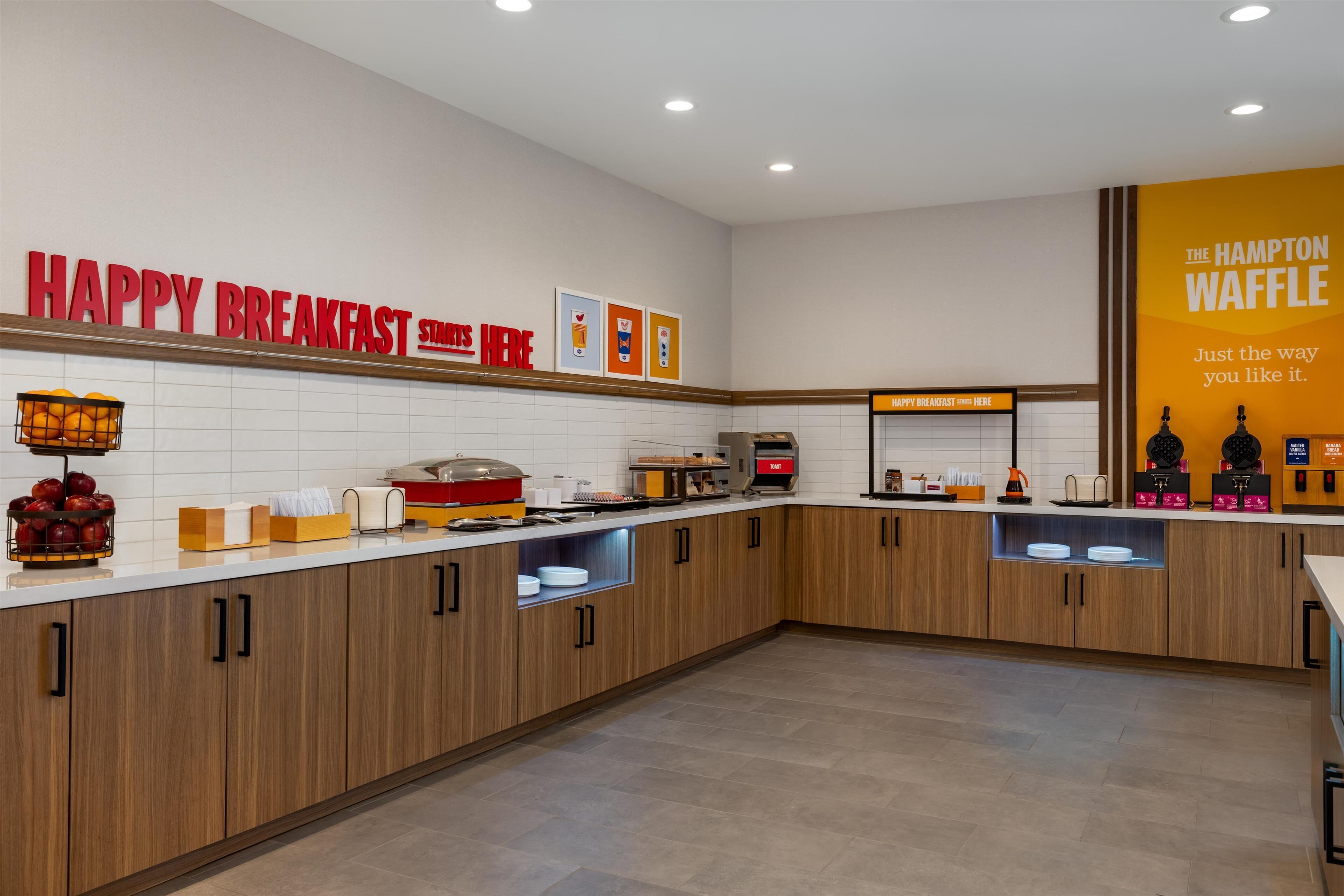 Un'area colazione moderna con un cartello 'Happy Breakfast', un cartello 'Hampton Waffle' e una macchina per il caffè.