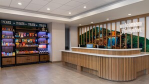 A hotel reception area with a 'Welcome to Hampton' sign, a 'Treats' section with snacks, and a wooden reception desk.