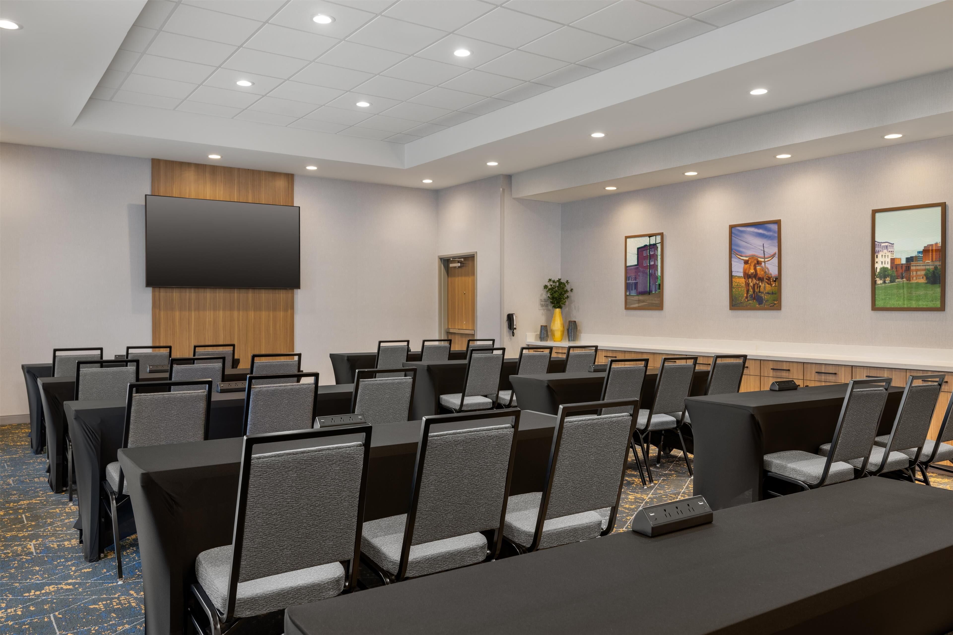 A conference room with rows of tables and chairs, a flat-screen TV, and framed pictures on the wall.