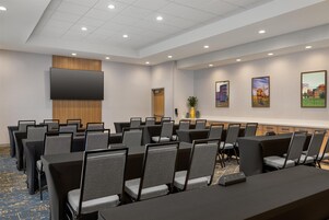 Un salón de conferencias con filas de mesas y sillas, un televisor de pantalla plana y cuadros en la pared.