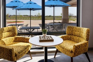 Dos sillones estampados enfrentados, con una mesita y una planta en maceta entre ellos, con vista a un patio con sombrillas azules y asientos al aire libre.