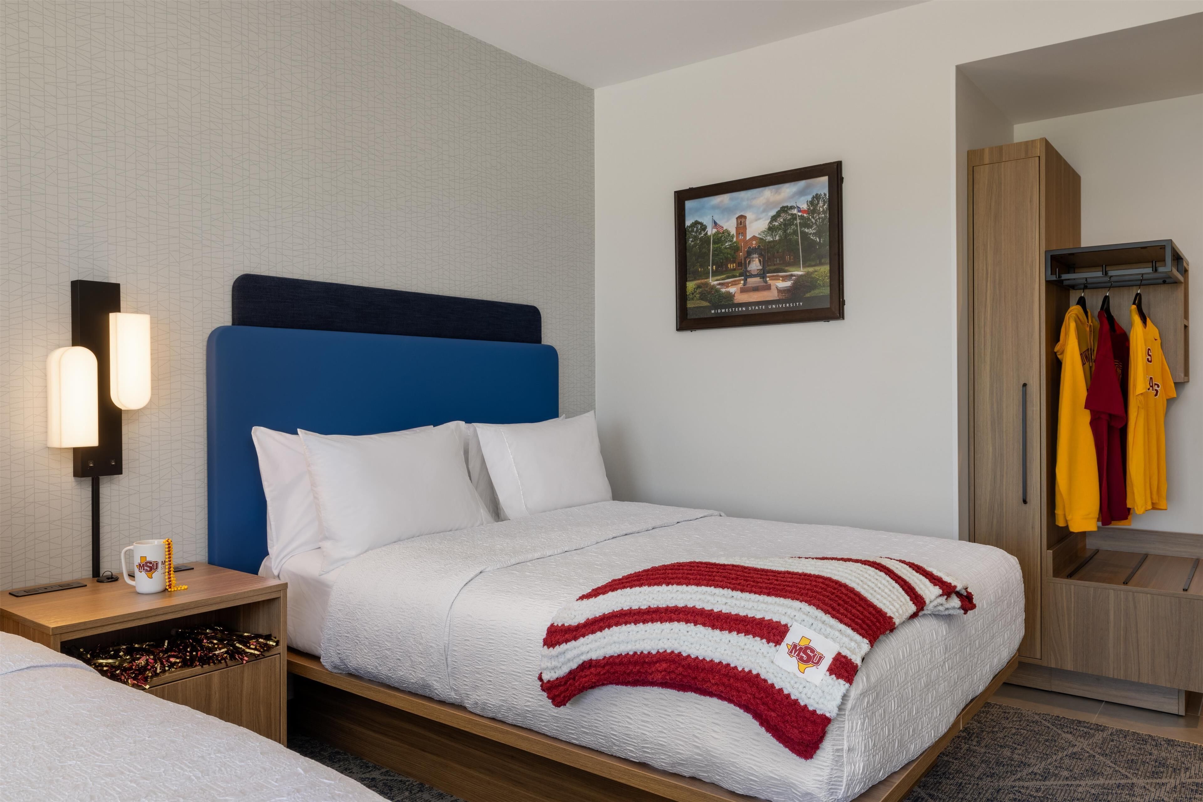 Habitación de hotel con cama, mesita de noche con lámpara, un cuadro en la pared y un armario con ropa colgada.