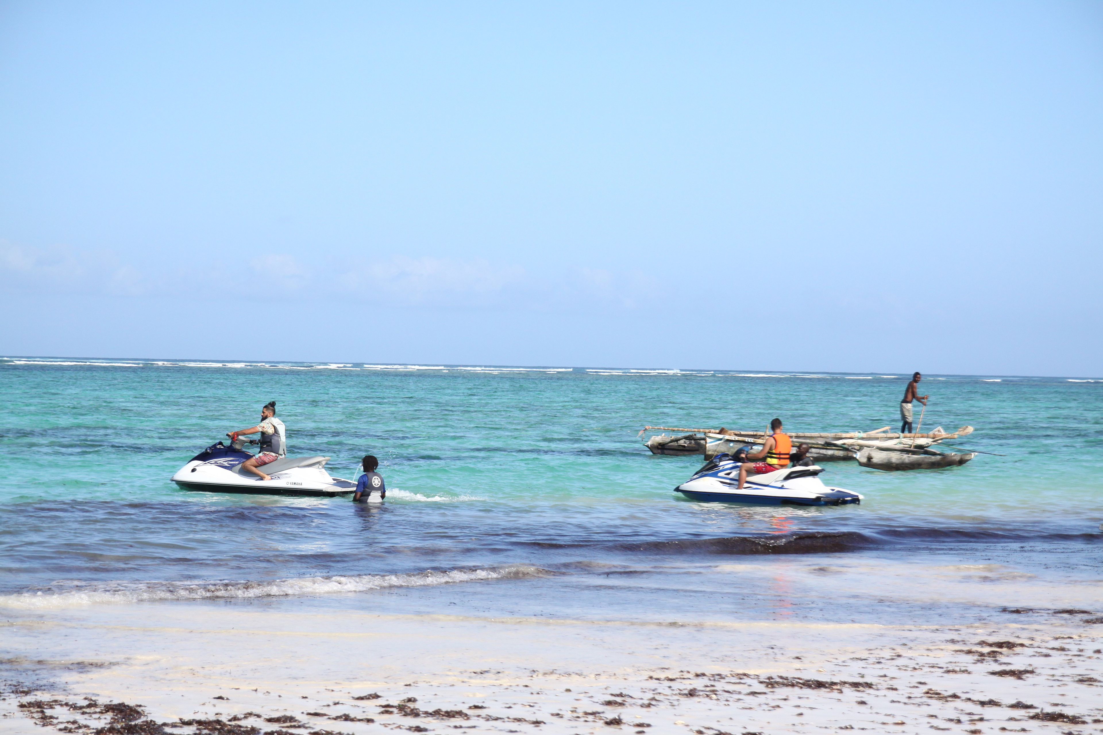 Escena de playa con motos acuáticas y pequeñas embarcaciones en el agua.