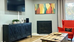 Una sala de estar moderna con un sofá de cuero negro, una mesa de centro de madera, un sillón rojo, un mueble de entretenimiento negro, un televisor de pantalla plana y un cuadro abstracto colorido.