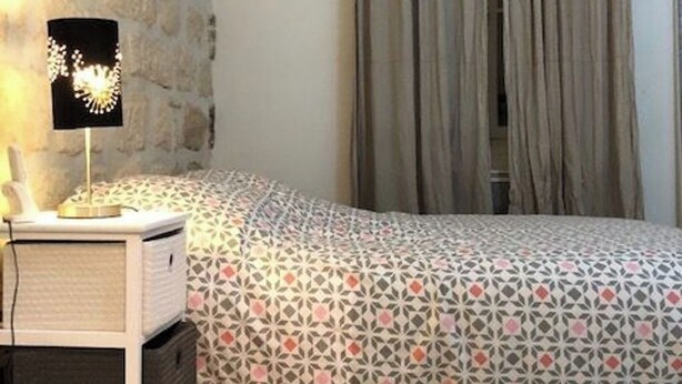 Une chambre à coucher avec une couvre-lit à motifs, une lampe de chevet, une petite armoire blanche et un mur en pierre.