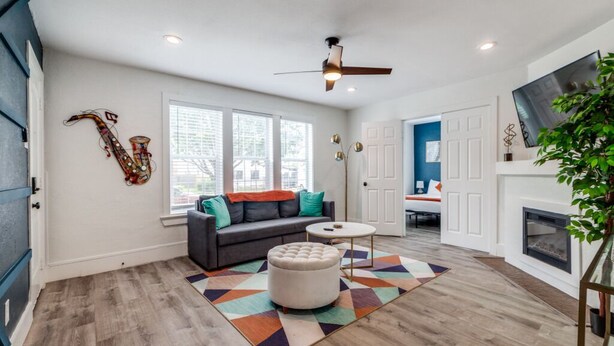 A spacious living room with a gray sofa, a round ottoman, and a colorful rug. There is a ceiling fan, a fireplace, and a large window with blinds. - Charming 6-bedroom house with AC in Houston (Houston)