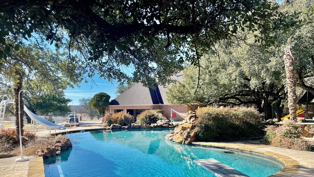 A swimming pool with a slide, surrounded by trees and a building with a sloped roof.