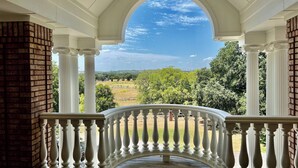 A covered porch with white balustrades and columns, offering a view of a green landscape and trees.