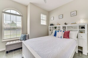 Une chambre à coucher avec un grand lit, un banc, une commode blanche et deux fenêtres équipées de stores.