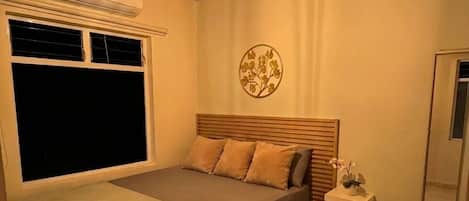 A bedroom with a bed, a bedside table, a wall-mounted air conditioner, a window with blinds, and a ceiling fan.