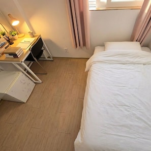Una habitación pequeña y ordenada, con una cama, un escritorio con lámpara y una mesita con una planta.