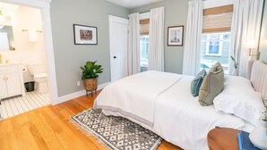 Un dormitorio con cama, mesita de noche, ventana con cortinas, lámpara de pared y puerta que da a otra habitación.