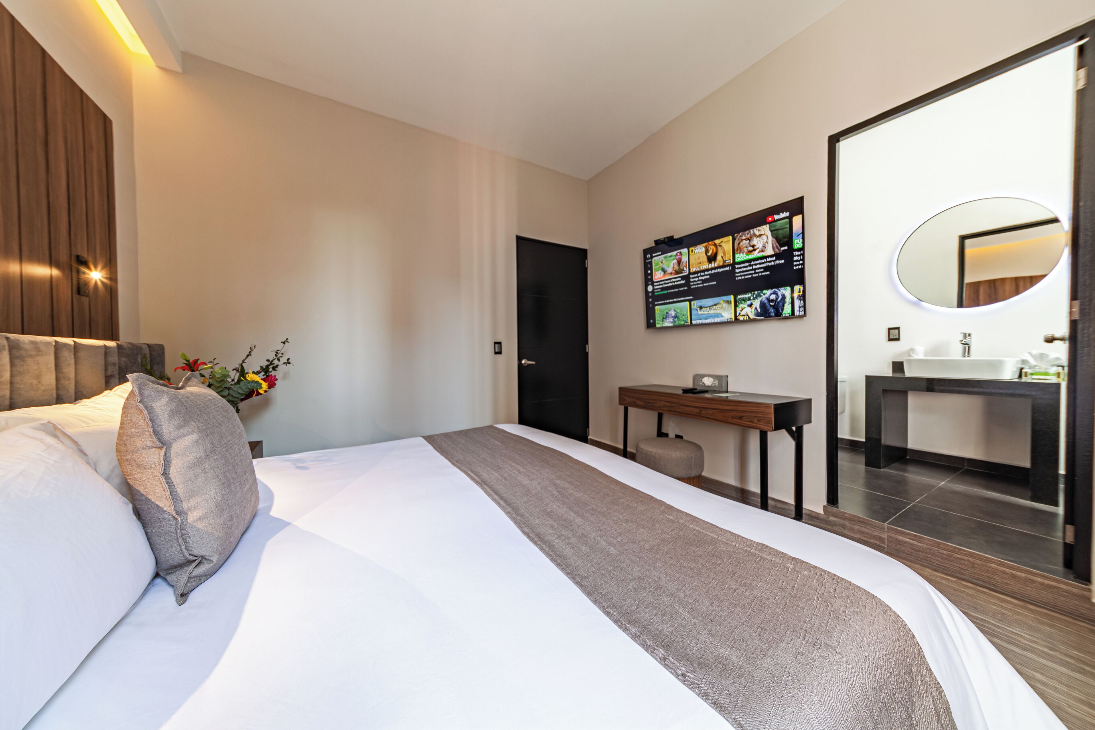 A modern hotel room with a large bed, a desk, a flat-screen TV mounted on the wall, and a bathroom with a large mirror and sink.