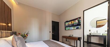A modern hotel room with a large bed, a desk, a flat-screen TV mounted on the wall, and a bathroom with a large mirror and sink.