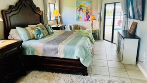 Un dormitorio con una cama grande, un ventilador de techo, un cuadro de palmeras y vistas al exterior.