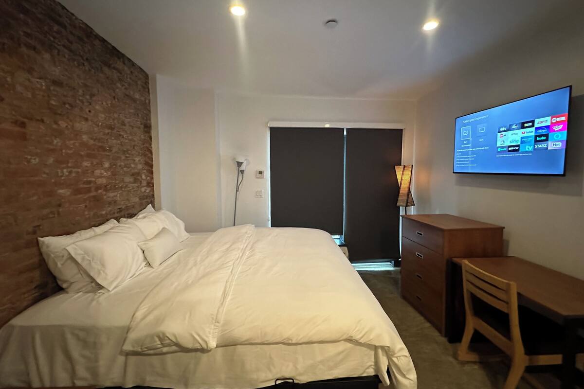 A hotel room with a brick wall, a bed with white bedding, a desk, and a TV displaying channel options.