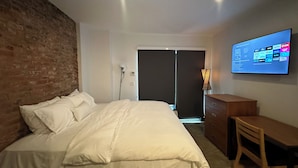 A hotel room with a brick wall, a bed with white bedding, a desk, and a TV displaying channel options.