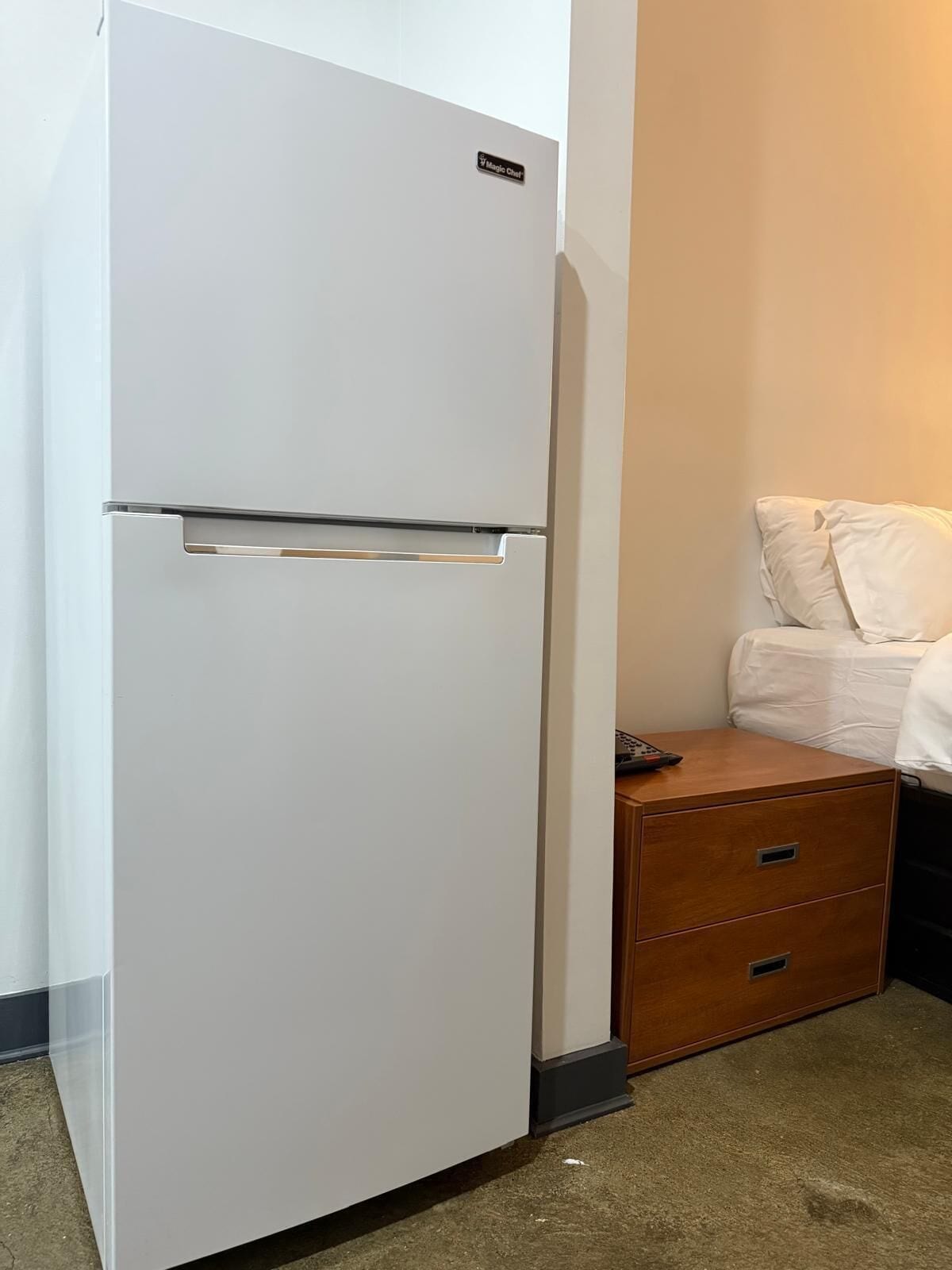 A white refrigerator in a hotel room with a wooden nightstand and a bed with white bedding.