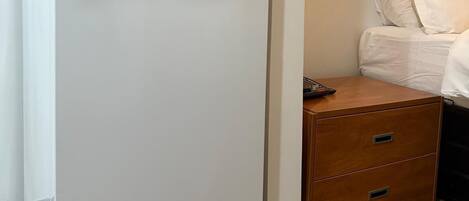 A white refrigerator in a hotel room with a wooden nightstand and a bed with white bedding.