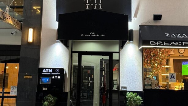 The exterior of the Tribeca Hotel, featuring a black awning with the hotel name, an ATM machine, and potted plants at the entrance.