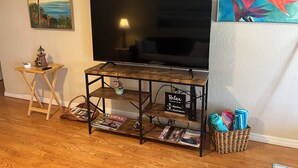 Una sala de estar con un televisor sobre un mueble de madera, una mesita con una lámpara y una cesta con ropa doblada.