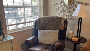 A room with a brown leather armchair, a patterned throw blanket, a round mirror on the wall, a lamp, and a small table with decorative items.