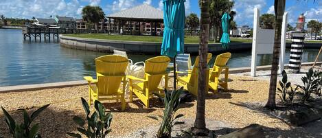 A lakeside seating area with yellow Adirondack chairs and blue umbrellas, surrounded by pebbles and plants.