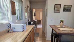 Une cuisine étroite avec des meubles de rangement en bois, un plan de travail et une poubelle. On y trouve deux fenêtres équipées de stores, un réfrigérateur et un four à micro-ondes.