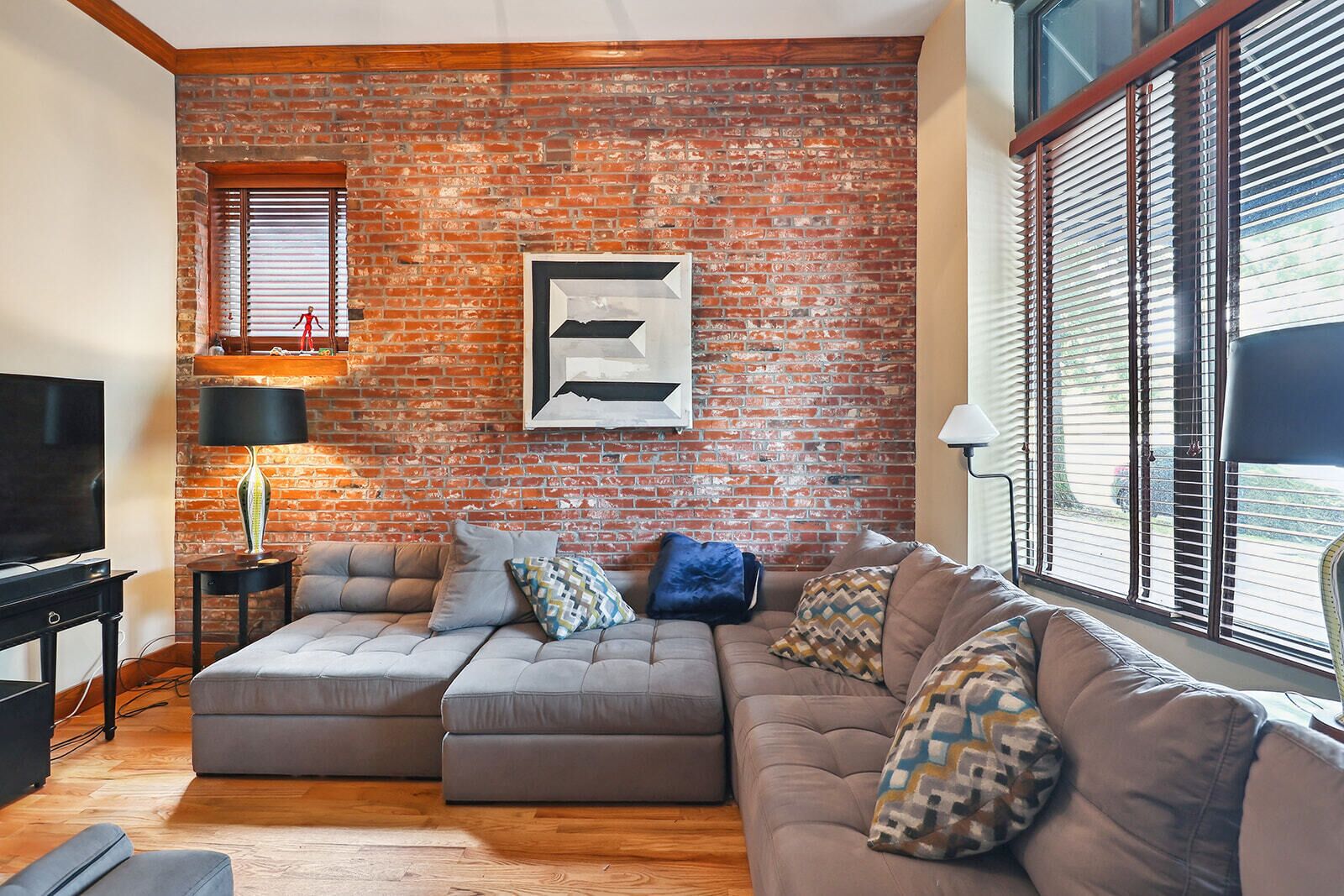 A living room with a brick wall, a sectional sofa, a television, and a window with blinds.