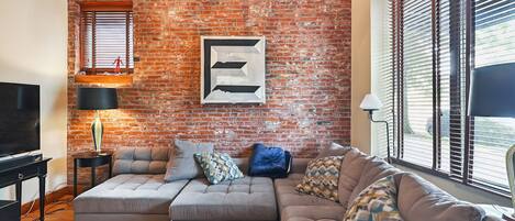 A living room with a brick wall, a sectional sofa, a television, and a window with blinds.