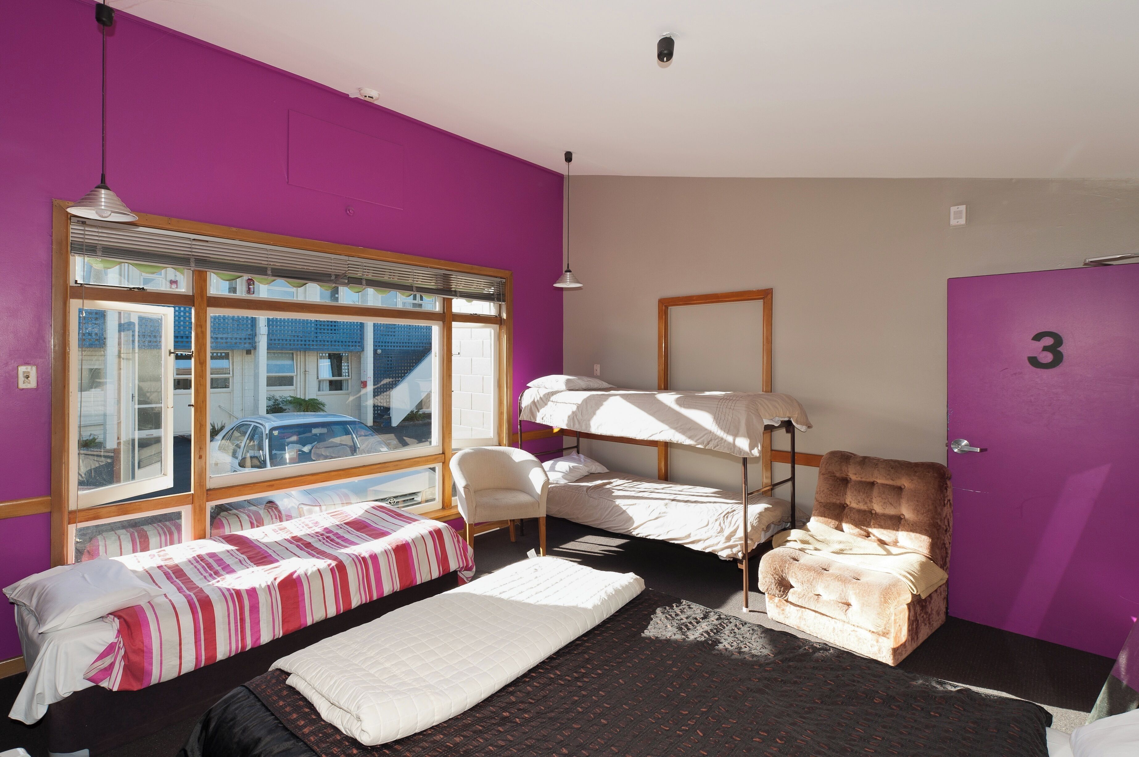 Une chambre avec un lit superposé, une chaise, un bureau et une fenêtre donnant sur une voiture et un immeuble.