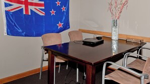 Un salón de conferencias con una mesa, sillas, una computadora portátil, un florero con ramas y una bandera.