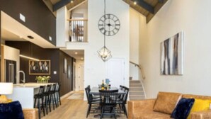 A spacious living area with a high ceiling, a dining table with chairs, and a large clock on the wall. - The Stella, minutes to the village/free shuttle (Mont-Tremblant)