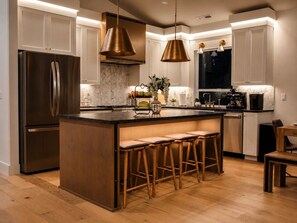 A modern kitchen with a central island, stainless steel appliances, pendant lighting, and wooden bar stools.
