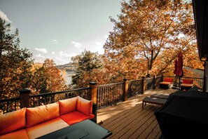 Una terraza de madera con mobiliario exterior, vista a un lago y follaje otoñal.