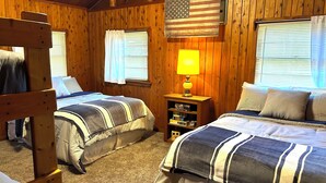 A cabin-style room with two beds, wooden walls, a small table with a lamp, and an American flag.