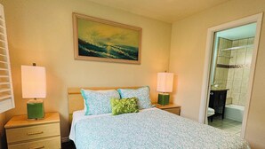 Ein Schlafzimmer mit einem großen Bett, zwei Nachttischen inklusive Lampen, einem Deckenventilator, einem Gemälde an der Wand und einem durch eine offene Tür einsehbaren Badezimmer.