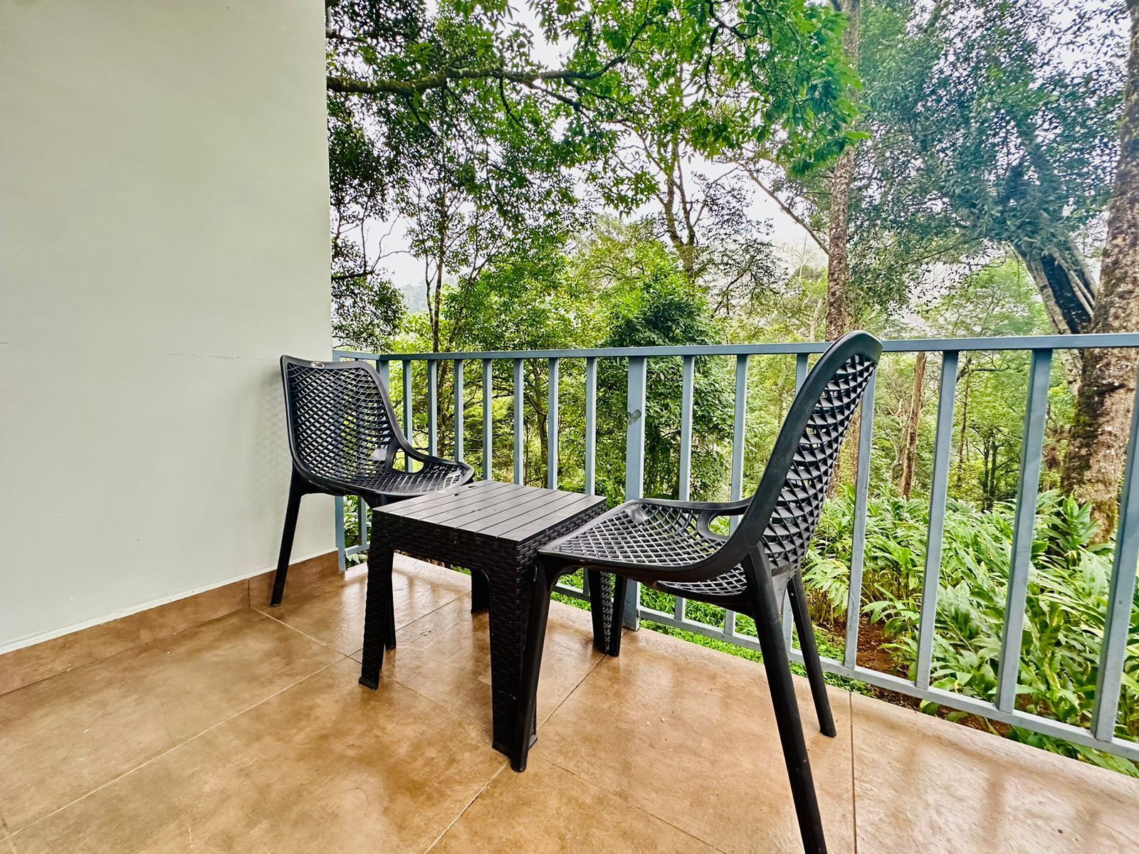A balcony with two chairs and a table, surrounded by a metal railing, overlooking a lush green landscape.