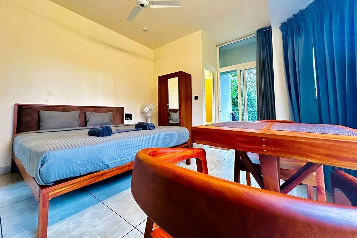 Ein Schlafzimmer mit einem Bett, einem Holztisch, einem Stuhl, einem Deckenventilator und einem Fenster mit Vorhängen.