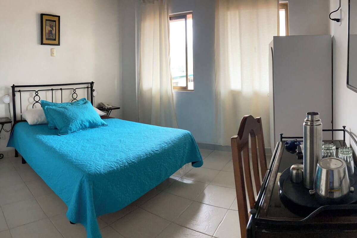 A bedroom with a bed, a bedside table, a chair, a desk with a kettle, and a refrigerator.