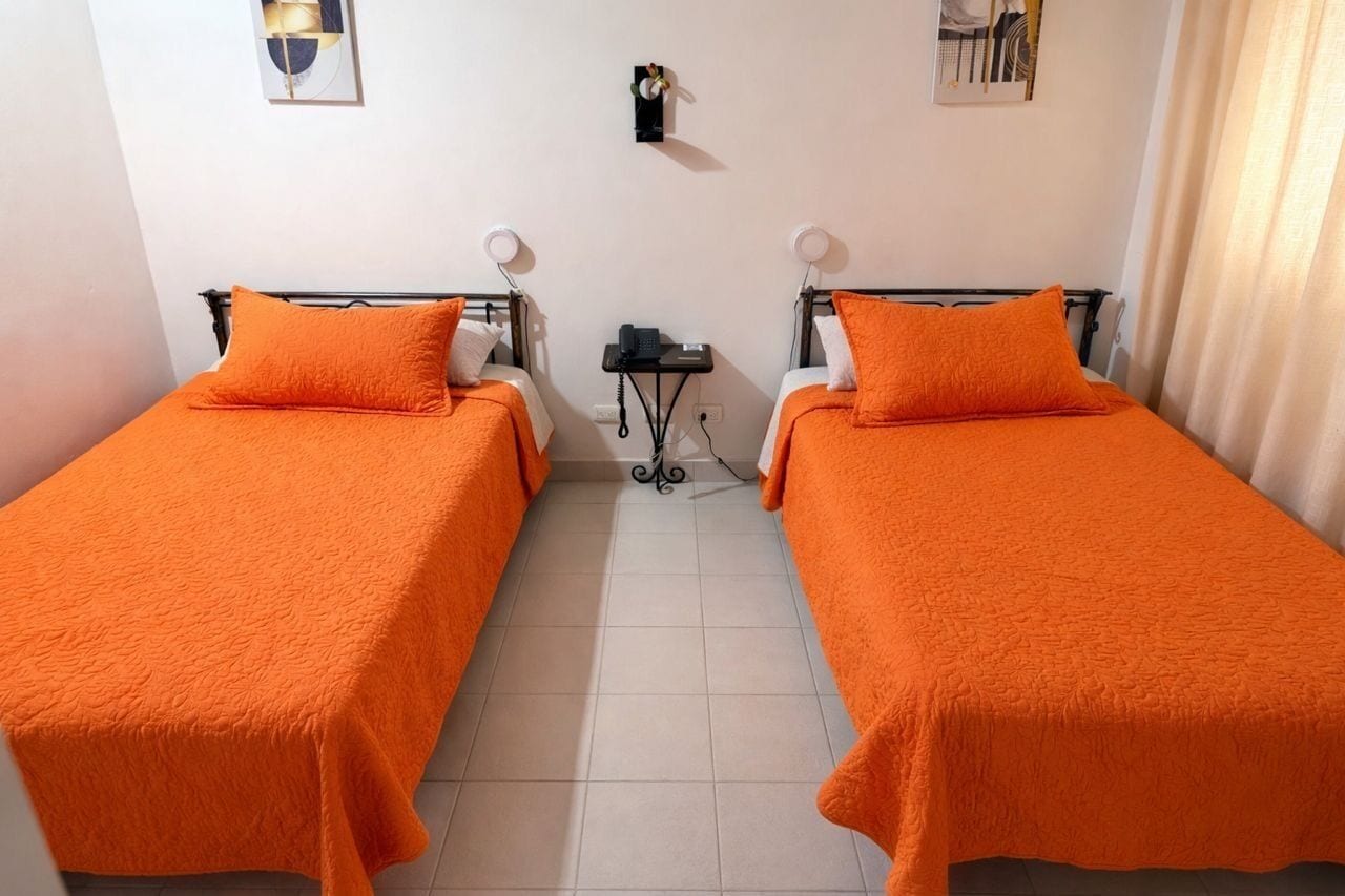 A room with two beds, each with an orange bedspread and pillow, a small table with a phone, and a wall-mounted light fixture.