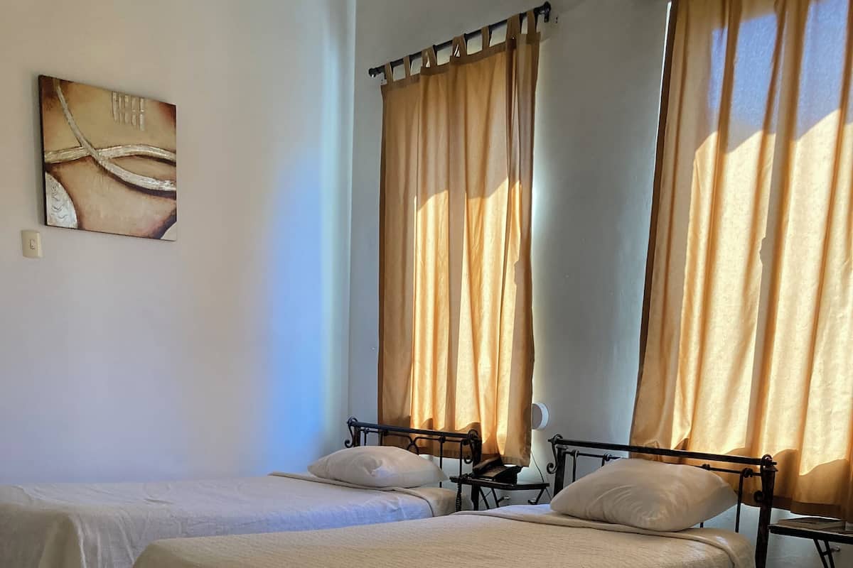 A hotel room with two single beds, beige bedding, a wall-mounted artwork, and curtains on the windows.