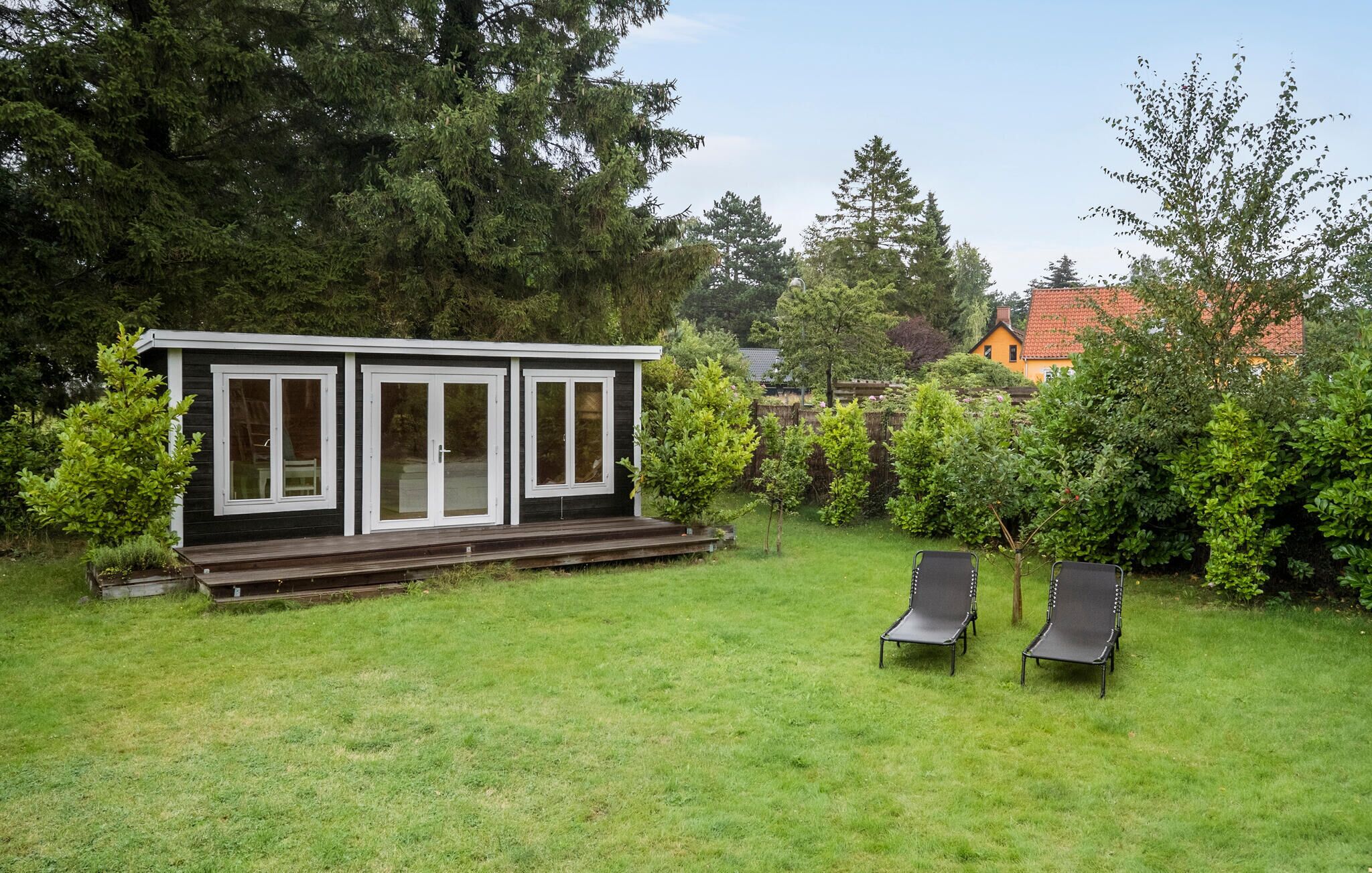 A small, modern cabin with a deck and two lounge chairs in a lush backyard.