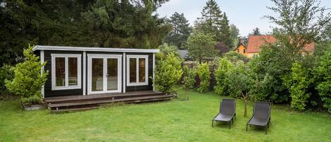 A small, modern cabin with a deck and two lounge chairs in a lush backyard.