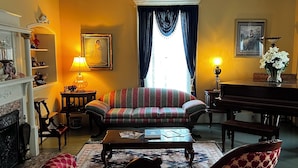 Un salon comprenant un canapé, un piano, une cheminée et une grande fenêtre avec des rideaux.