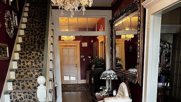 Un magnifique escalier, agrémenté d’une moquette à motifs floraux, d’un lustre et d’un miroir encadré d’un décor ouvragé.
