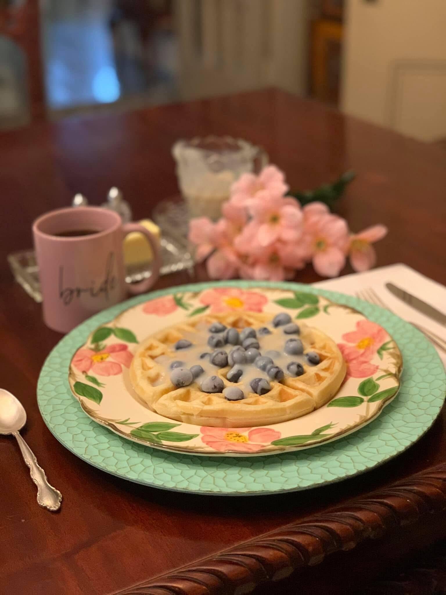 Un plato de gofres con arándanos sobre un plato estampado con flores, una taza rosa con la etiqueta 'novia' y flores rosadas sobre una mesa.