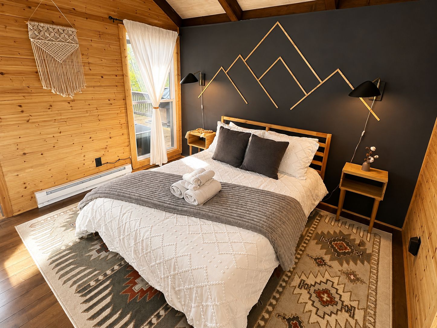 Een slaapkamer met een houten plafond, een bed met wit en grijs beddengoed, een kamerbreed tapijt met een patroon en een geometrische lamp aan de muur.