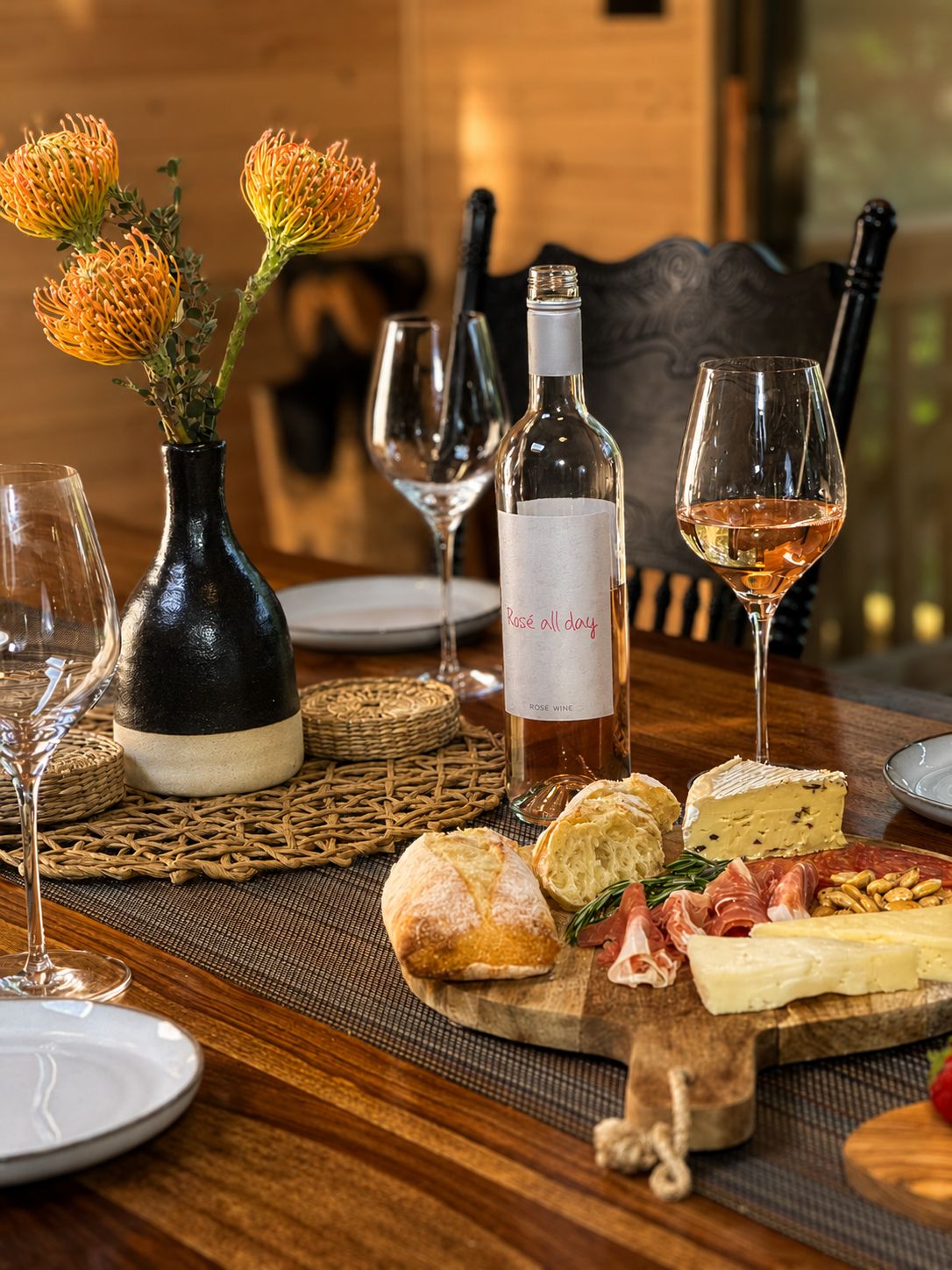 Ein Esstisch mit einer Roséweinflasche, Gläsern, einer Käseplatte, Brot und Blumen.