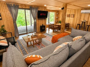 A cozy living room with a sofa, a coffee table, and a fireplace. A television is mounted on the wall, and there is a large window with curtains. - CasaSOL: Stylish Mountain Cabin with Game Room, Trails and Fire Pit Nights (Basye)